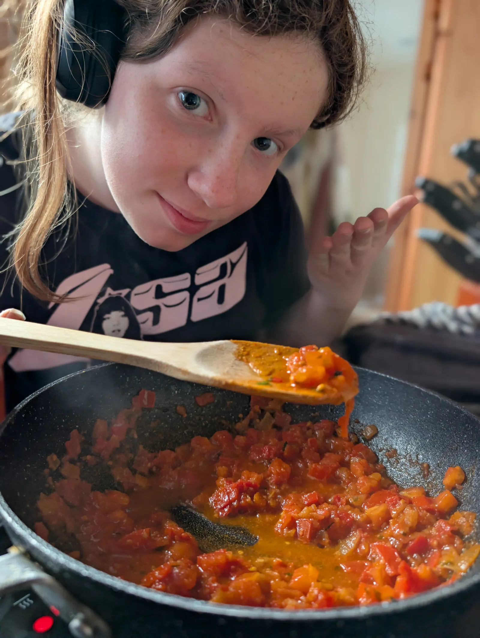 Ava at the hob, headphones on, lifting a wooden spoon over a pan of chopped tomato and onion curry sauce