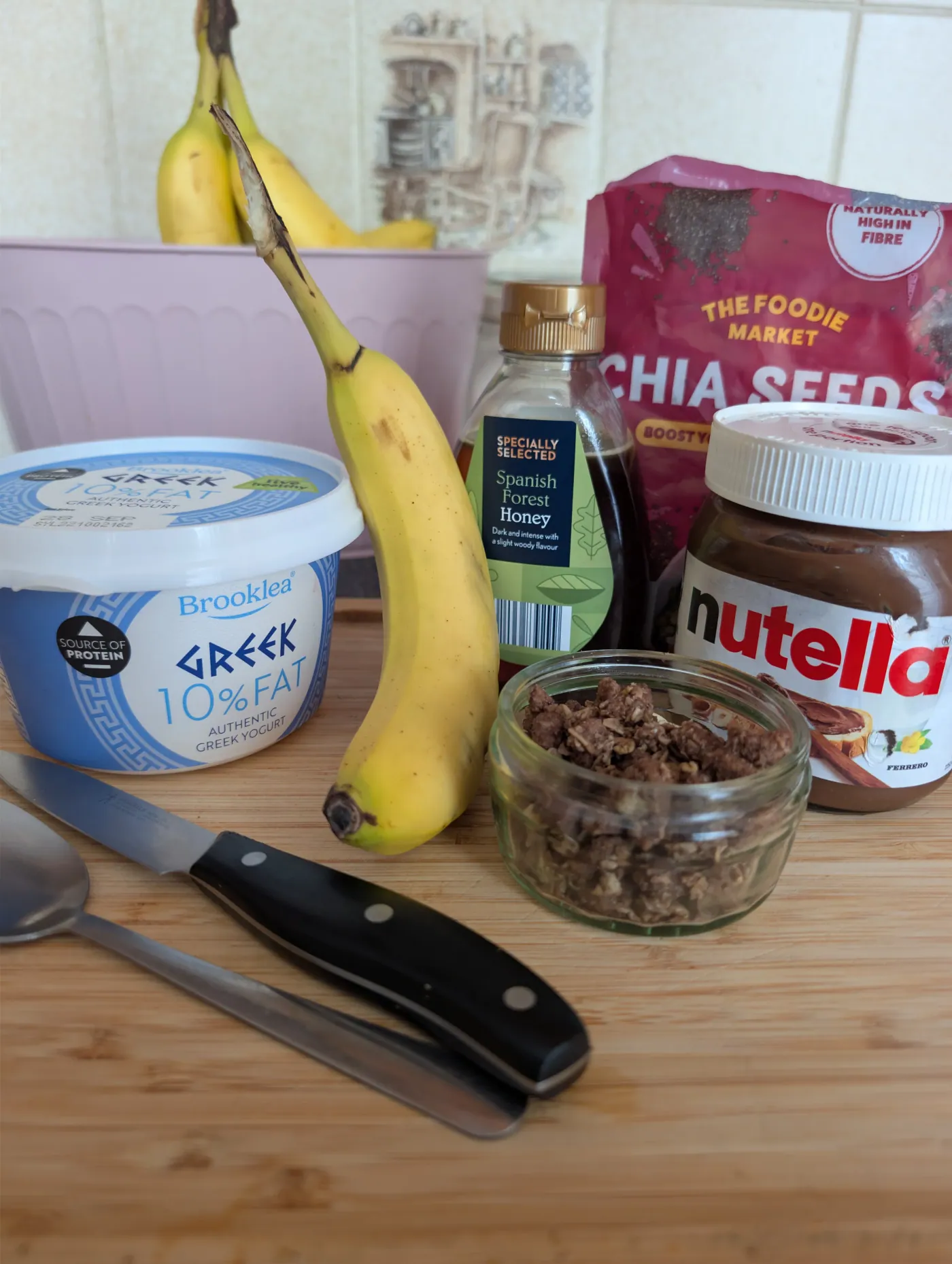 Greek yoghurt, a banana, honey, chia seeds, Nutella and a jar of granola laid out on a wooden board with a knife and spoon