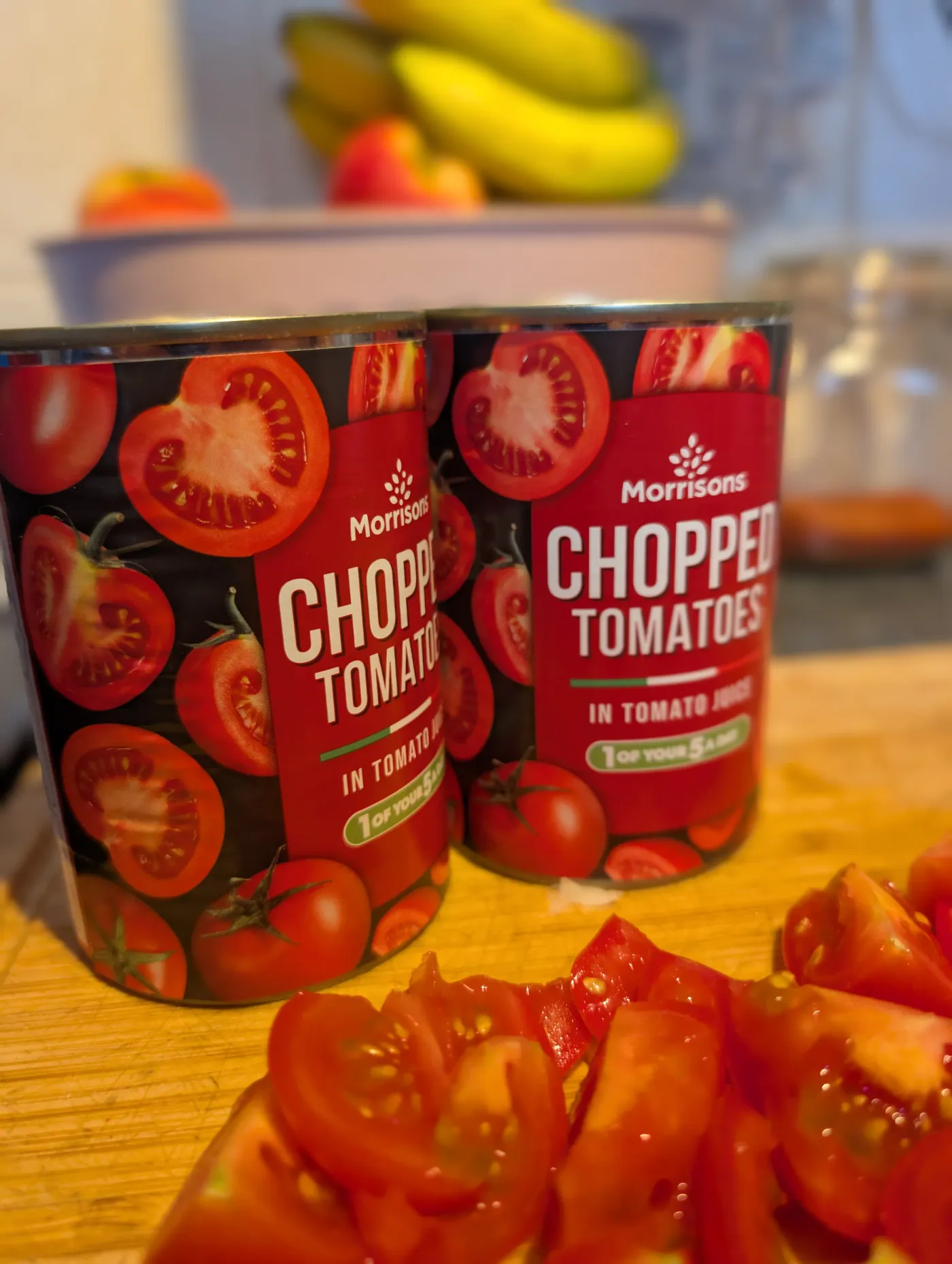 Two tins of chopped tomatoes on a wooden board beside a pile of freshly chopped tomatoes